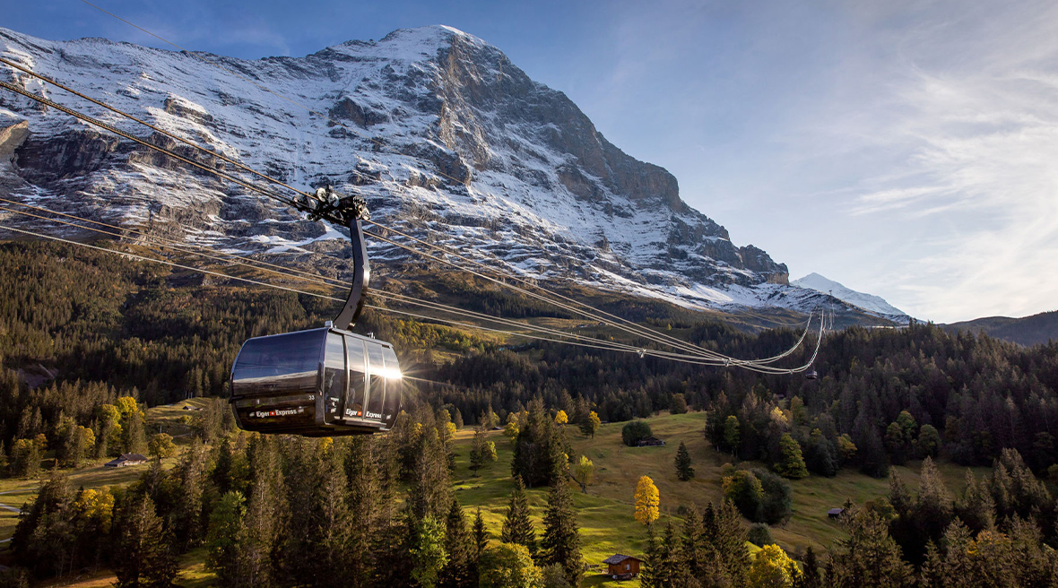A brand new, sleek grey cable car hangs off a three-cabled line against a backdrop of grassy valley and snowy peaks